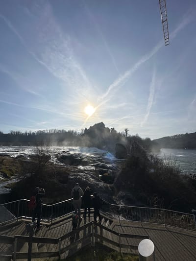 This image depicts a panoramic view of the Rhine Falls in Neuhausen am Rheinfall, Switzerland, captured on a brightly lit day. The sun, a prominent luminous disc with visible rays, is positioned in the upper-center sky, casting a strong backlight over the entire scene. The sky is light blue with multiple white contrails crisscrossing, and a segment of a yellow construction crane boom is visible in the upper right corner.
In the mid-ground, the powerful Rhine Falls cascade extensively over rocky terrain, generating significant mist that partially obscures the historical Schloss Laufen (Laufen Castle), situated on a cliff overlooking the falls. The river continues downstream to the right. The surrounding landscape features bare trees and bushes, suggesting a cooler season.
In the immediate foreground, a multi-level wooden viewing platform with dark metal railings is visible. Six individuals are present on this platform, predominantly viewed from behind and silhouetted by the sun. One person on the far left, wearing a dark coat and a red backpack, is holding a device up, likely a camera or phone, directed towards the falls. To their right, another individual wears a light-colored backpack, standing near a small child. The people are observing the falls. Long, distinct shadows are cast by the railings and individuals across the wooden platform. A cylindrical metal post with a circular, reflective top is situated on the lower right section of the platform.