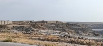 The image shows a large, excavated area, likely a quarry or construction site, in Margilan City, Uzbekistan. The foreground features dry, golden grass and a paved surface, possibly a road. A fence made of precast concrete segments runs horizontally across the middle ground, behind which several industrial buildings with light-colored roofs are visible. These buildings appear to be long, rectangular structures, suggesting warehouses or manufacturing facilities. The terrain is rugged and uneven, with layered rock formations and piles of soil and debris. In the distance, a hazy horizon line suggests more flat land with sparse vegetation and utility poles. The sky is clear and pale, indicating daytime. There are no people or animals visible in the image. The scene suggests an industrial or commercial development in a semi-arid environment.