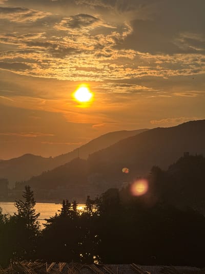 The image captures a sunset over a coastal city, likely in the vicinity of Vrijesno, Montenegro. The sun, a bright, glowing orb, is positioned in the upper left quadrant of the sky, casting a warm, golden light across the scene. Wispy clouds, illuminated by the setting sun, stretch across the sky, creating a dramatic and colorful display.
In the foreground, silhouetted trees and foliage obscure parts of the view. A body of water, possibly the Adriatic Sea, is visible in the middle ground, reflecting the golden hues of the sunset. Beyond the water, a range of hills or mountains rise, their slopes dotted with buildings and structures, suggesting an urban or semi-urban environment. A prominent tower or antenna stands on one of the hilltops.
In the sky, a small, colorful parachute, likely from a paraglider or parasailer, is visible in the upper right quadrant, adding a touch of human activity to the natural landscape. The overall impression is one of serene beauty and tranquility, with the vibrant colors of the sunset dominating the scene.