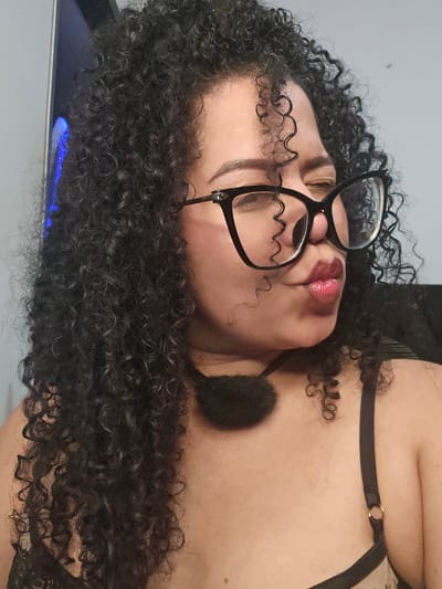 A woman with dark curly hair and glasses is lying down, facing upward. She is wearing black lingerie. Her lips are glossy and red. A black, round object is positioned near her neck. The image provides no information about the location in the city of Perímetro Urbano Cúcuta, Colombia.
