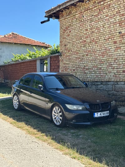 The image shows a black BMW sedan parked on a grassy area next to a gravel path. The car has a Bulgarian license plate that reads "T 4744 AM". Behind the car is a brick wall of a building, and further in the background, there is a tiled roof of another building and some greenery. The sky is clear and blue, indicating a sunny day. There are no people visible in the image. The scene appears to be in a rural or suburban area. Based on the license plate and the general architecture, the location is likely Konop, Bulgaria.
