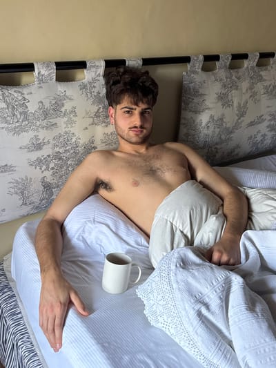 A young adult male is depicted reclining in a bed within a room located in Montepulciano, Italy. The subject, with dark curly hair, a mustache, and dark eyes, gazes directly forward. His upper torso is bare, displaying chest and armpit hair, a thin necklace, and a stud earring in his left ear. He is positioned against a bedhead comprising a black metal frame and two rectangular fabric panels patterned with grayscale toile de Jouy pastoral scenes. The bed is made with white striped and plain pillows, a white striped fitted sheet, a light-colored duvet, and a white decorative blanket featuring intricate patterns. A plain white ceramic mug containing a dark liquid is situated on the sheet beside the subject's left arm. A plain, light-colored wall is visible in the background.