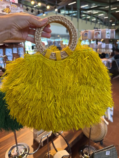A light-skinned human hand firmly grasps a circular, woven rattan handle of a handbag in an indoor retail setting located in the City of Panglao, Philippines. The handbag itself is predominantly bright yellow, constructed from dense, shaggy, natural fibers, likely raffia or straw, giving it a voluminous, fringed texture. The rattan handle, approximately 10-12 cm in diameter, is secured to the bag's body with yellow fabric strips. In the blurred background, various retail displays are visible, including a sign partially showing "Mango" with an image of mango slices, suggesting food or souvenir sales. Other woven items, including a similar textured bag in green, are present on metal display stands. A circular woven basket and a flat, woven rectangular item are also discernible on a dark surface in the lower right. A blurred individual is partially visible in the far background on the right. The environment is illuminated by warm overhead lights.