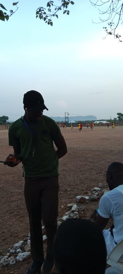 A coach stands on a reddish-brown field, observing a soccer practice in the distance. He wears a green t-shirt, brown pants, and a black cap, and has a lanyard around his neck. To the right, a person in a white shirt sits in a chair, partially visible. Rocks are scattered on the ground. In the background, a few utility poles are visible, and a mountain range is present on the horizon beneath a partly cloudy sky. Branches with leaves frame the top corners of the image.