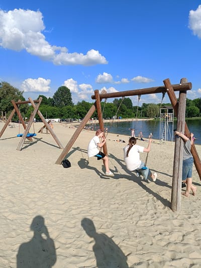 On a sandy beach under a bright blue sky with scattered white clouds, two young females are on swings. The female on the left, wearing a white t-shirt and black shorts, is on a swing with a blue seat. The female on the right, also in a white t-shirt and blue shorts, is on another swing with a blue seat. Behind them, other people are visible on the beach and in the water of a lake. A lifeguard tower stands near the water. Wooden swing sets are prominent in the foreground. The scene is set at a beach in Gubkin, Russia.