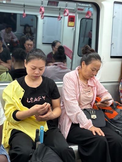 In 龙溪街道, China, inside a subway car, two women are seated. The woman on the left wears a black t-shirt with the word "LEAFY" in pink lettering and a bright yellow jacket over her shoulders. She is looking down at her phone, holding it in her hands. To her right, another woman wears a light pink jacket over a pink t-shirt. She is also looking down at her phone, holding it in her right hand. Both women are seated on a silver bench. Between them, a black handbag with a gold clasp shaped like rabbit ears rests on the seat. A bright orange and black backpack is visible behind the woman in pink. Other passengers are visible in the background, some looking at their phones, others looking out windows or around the car. Handrails are visible overhead.