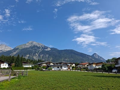 The image depicts a residential area in Münster, Austria, nestled at the base of imposing mountains. In the foreground, a lush green field is dotted with yellow wildflowers. A rustic wooden fence with metal inserts separates the field from a paved walkway. Behind the fence, several houses with pitched roofs, some featuring solar panels and balconies with flower boxes, are visible. A dense line of evergreen trees forms a backdrop to the houses, with the rugged, tree-covered mountains rising dramatically behind them. The sky above is a brilliant blue, adorned with scattered white, wispy clouds. A white car is parked on the road in front of one of the houses.