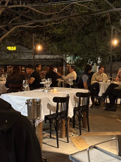 The image depicts an outdoor dining area at night, specifically identified as El Chacal restaurant, located at Calle Ocampo 25, Zona Centro, Guadalajara, Jalisco, Mexico. Approximately ten individuals are visible, seated at various tables.

In the immediate foreground, the back of a person wearing a black hoodie is partially visible to the left. A square table, covered with a white tablecloth, features wine glasses and a small lit candle, with a silver metal ice bucket on a stand positioned next to it. Black wooden chairs with slatted backs are pulled up to the tables.

In the mid-ground, a group of five people is seated at two adjacent tables. This group includes a female with dark hair seen from behind, two males engaged in conversation (one in a black polo shirt, the other in a black shirt), another female with dark hair seen from behind, and an older male in a light-colored shirt actively gesturing. Further right, a male in a dark vest is seated opposite another male in a beige shirt who is looking down at a mobile device. Partial views of additional patrons and their legs, one with crossed legs in dark pants and shoes, are also present.

Overhead, a metal trellis structure supports warm incandescent light bulbs, interspersed with dense tree branches and foliage, creating a canopy. In the background, a building with a corrugated metal roof is visible, displaying a neon sign that reads "El Chacal." The flooring consists of concrete, patterned tiles, and integrated wooden planks. Patrons are engaged in dining, conversation, and individual activity.