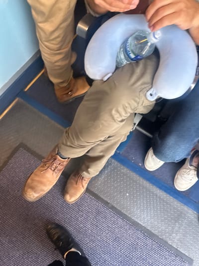 In an indoor environment, possibly an airplane cabin, a person's legs are visible, wearing tan trousers and brown lace-up boots. Another person's legs are seen wearing blue jeans and white sneakers. A light blue neck pillow with a water bottle is placed on the lap of the person in tan trousers. The floor is covered in a blue patterned carpet. The location in the city of Uttlesford, United Kingdom, is not discernible from the image.