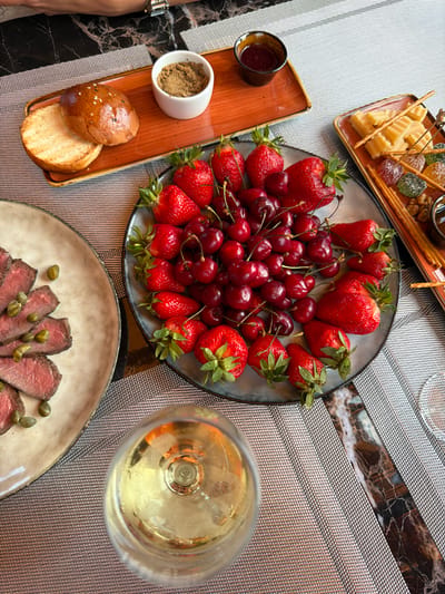 A high-angle, close-up shot displays a sophisticated dining spread, possibly in a restaurant in Gorno-Altaysk, Russia, featuring a variety of dishes and drinks. The table is set with a textured placemat overlaying a dark, marble-patterned surface.

Dominating the center is a circular blue-gray plate filled with fresh strawberries and dark cherries, their vibrant colors contrasting with the plate. To the left, a beige, oval-shaped plate holds thinly sliced, rare roast beef topped with capers. In the foreground, a clear wine glass filled with a pale yellow liquid, likely white wine, rests on the placemat.

In the upper left quadrant, a rectangular orange tray holds a split bun with sesame seeds and a small white ramekin filled with a brown spread, possibly a dip or paté. Adjacent to this, another orange tray contains a small dark dish of red jam or sauce. To the right, a rectangular platter presents cubes of light yellow cheese, breadsticks, and several colorful, translucent candies, possibly jelly or fruit jellies, along with nuts.

A person's arm, wearing a watch with a metallic band, is visible in the top left corner, suggesting they are seated at the table. The lighting is bright and even, highlighting the textures and colors of the food and tableware. The overall impression is one of a leisurely and upscale meal.