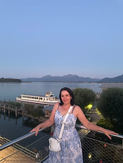 A dark-haired woman in a blue and white patterned dress and a white crossbody bag stands on a metal walkway, leaning on the railing. The walkway overlooks a body of water, where a large white boat with green trim is docked. In the background, a line of trees separates the water from mountains under a twilight sky. The scene is set in Prien am Chiemsee, Germany, possibly at the harbor.