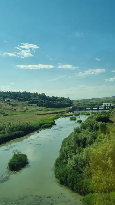 The image captures a serene landscape with a winding river flowing through lush green hills under a bright blue sky dotted with scattered white clouds. The river, with its calm, greenish waters, is bordered by dense vegetation and reeds. In the background, a bridge structure, possibly a dam or a causeway, spans across the river, with several upright pillars visible. Rolling hills covered in trees and grass extend into the distance, creating a picturesque rural scene. The overall impression is one of natural beauty and tranquility, with no human figures or specific urban elements readily apparent. The specific location in the city of Șcheia, Romania, is not visually identifiable from the image itself without external information.