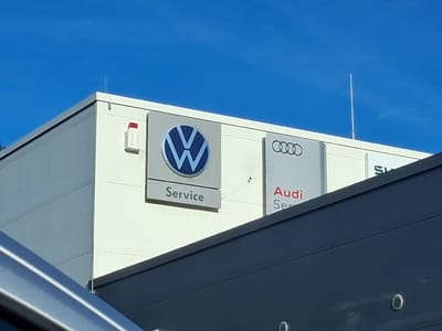 The image shows the exterior of a building with signage for Volkswagen, Audi, and Skoda service centers. The Volkswagen sign features the circular VW logo and the word "Service" below it. The Audi sign displays the four interconnected rings logo and the word "Audi" above "Service". The Skoda sign is partially visible with the "SK" of the logo. The building has a light-colored exterior wall and a darker colored overhang. The sky above is clear and blue. The scene is located in Vilshofen an der Donau, Germany.