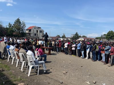 A large group of people are gathered in an open area, likely a public square or a parking lot, in the city of Goma, Democratic Republic of Congo. A man is speaking from a makeshift stage, positioned on a raised platform and made of wood planks. He is wearing a dark-colored suit jacket. The crowd is seated on white plastic chairs facing the speaker, with additional people standing behind them. The crowd is predominantly male, with a few women visible. There are trees and buildings in the background, and the sky is clear and blue. The ground is covered in gravel and dirt. The scene suggests a public address or a community gathering.