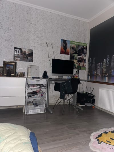 A cluttered room features a desk with a computer and a black chair. Posters and a city skyline print adorn the wall above. A white storage unit with drawers sits near the desk, topped with a wire basket filled with items. A rug with a smiley face is visible in the upper right corner, and a bed with blue accents is in the lower right corner. The room has light-colored patterned wallpaper and gray flooring.