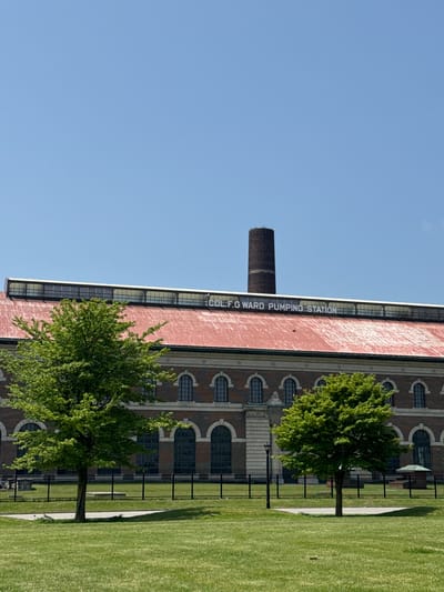 The image shows a brick building with arched windows and a smokestack. The building has text on its side that reads "D.F.C. WARD PUMPING STATION". There are trees in front of the building and a grassy lawn with a fence. The sky is clear and blue, suggesting a sunny day. The scene appears to be a historical industrial site, likely in Buffalo, United States of America, given the location context. There are no people visible in the image, and the overall mood is calm and serene, highlighting the architecture and natural elements. The time of day appears to be daytime, possibly late morning or afternoon, due to the bright sunlight casting shadows. The architectural style of the building suggests it is an older structure, possibly from the late 19th or early 20th century. The presence of the smokestack further reinforces its industrial past. The neatly kept lawn and trees indicate that the site is either preserved or repurposed for public use, such as a park or museum.