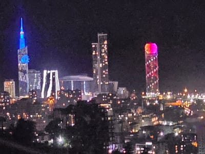 The media file captures a vibrant night-time cityscape, likely Batumi, Georgia, as viewed from a distance, possibly from a location like ქვედა სამება. The scene is dominated by the artificial glow of numerous buildings against a dark, clear night sky.
On the far left, a prominent, slender skyscraper stands out, illuminated predominantly in bright blue light, with a distinctive spire at its peak. A unique, intricate structure, possibly spherical or rotating, is also visible around its upper section, glowing blue. This building is identifiable as the Alphabet Tower. To its right, another cluster of high-rise buildings features numerous internal lights. Further in the background, a large, flat, disc-like structure supported by columns is faintly visible.
On the right side of the frame, another tall, slender building is brightly illuminated in striking pink and red hues. Its very top features a large, glowing spherical structure, likely indicating a hotel or entertainment complex. This is consistent with the Radisson Blu Hotel.
The mid-ground and background are densely packed with the scattered, warm lights of smaller buildings, streets, and urban infrastructure, creating a dynamic urban tapestry. The immediate foreground shows dark, silhouetted foliage or terrain, suggesting the photo was taken from an elevated vantage point.
The image conveys the lively atmosphere of a modern city after dark. There is no visible human activity or discernible legible text due to the distance and resolution, although the Alphabet Tower is known for displaying the Georgian alphabet. The time of day is clearly night, and the weather appears calm and clear.