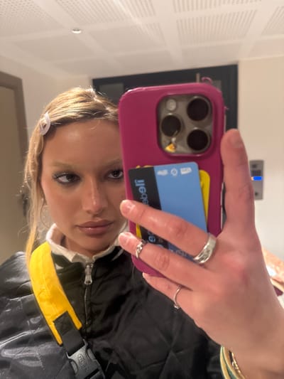 A person takes a selfie in front of a wall. They are holding a pink phone case with a blue credit card and a black transit card tucked into the back. The person has several rings on their fingers. They are wearing a black jacket with a yellow strap. The image is taken at an angle, and the person's face is tilted.