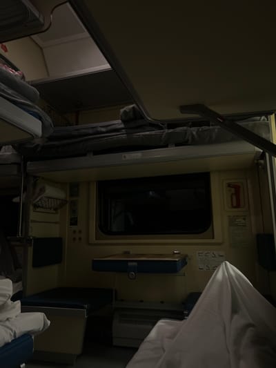 The image captures the interior of a dimly lit train sleeper compartment, oriented as if viewed from one end, looking towards the window. The overall ambiance suggests nighttime travel.

The scene is dominated by light cream-colored paneling typical of train cabins. A central rectangular window, dark with no visible exterior, confirms the low-light conditions. To the right of the window, a small shelf is visible, holding a metal basket containing a rolled white item, possibly a towel or blanket, and some small cutlery or utensils. A dark blue object, possibly a book or tablet, rests beside it. Above this shelf, there are electrical outlets, switches, and what appears to be a small light fixture.

Above the window, a red "P"-shaped handle, likely an emergency brake release or safety mechanism, is mounted on the wall. Adjacent to it are printed instructions in Cyrillic script, though unreadable, along with safety diagrams. Further right, a set of diagrams illustrates various configurations for the bunk beds, depicting stick figures in sitting and sleeping positions. Dark grey curtains are drawn or tied back to the left of the window.

Two bunk beds are present within the compartment. The upper bunk is partially visible in the top left corner, made with white bedding. The lower bunk is seen in the bottom right, also with white bedding and some blue accents, suggesting a comfortable sleeping arrangement. A ventilation grille is visible on the upper right side of the compartment wall. To the far left, an open doorway reveals a glimpse into another dimly lit area, possibly a corridor or an adjoining space.

No people are visible in the compartment. The setting strongly suggests a Russian long-distance train, consistent with the provided location hint of Vodorazdel, Russia. The scene conveys a sense of quiet and rest during travel.