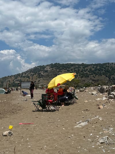 The image depicts a beach scene in Ortaca, Turkey. A group of people are situated under a yellow umbrella, seated in folding chairs. Surrounding them are beach accessories and personal belongings. Behind them is a rocky hillside. The sky above features a mix of blue and white clouds. The beach itself is composed of sand, rocks, and scattered shells.