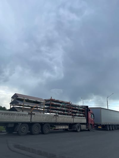 The image shows a semi-trailer truck carrying a load of what appear to be prefabricated building components in Бавыкино, Russia. The truck is positioned on an asphalt road under an overcast sky. The load is secured with straps and consists of multiple large, flat panels supported by a metal frame. Another truck, a semi-trailer type, is positioned behind the first one. Streetlights are visible in the background.