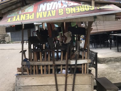 The image depicts a food stall in Rambot, Indonesia, under a makeshift awning advertising "GORENG & AYAM PENYET EKWAN." The stall is constructed of wood and contains a variety of tools, primarily hoes and axes, arranged within its structure. In the background, there is a street scene with buildings and an outdoor seating area with tables and chairs. The ground is paved, and the overall scene appears to be a typical urban setting in Rambot. There are no people visible in the immediate foreground.