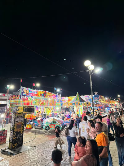 A lively carnival scene unfolds at night, illuminated by bright lights. People crowd around a colorful ride, with various characters like Spider-Man and Frozen adorned on its facade. A street lamp casts its glow, and power lines stretch across the dark sky. The foreground shows a platform with arcade games and a family observing the spectacle. The scene is vibrant with excitement and activity.