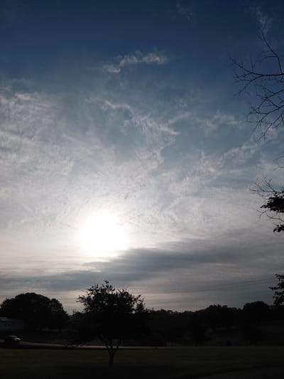 This is a low-angle, full-shot photograph of a landscape at dusk. The sky occupies the majority of the frame, featuring a gradient of dark blue at the top to lighter blue and white hues as it descends. wispy clouds are scattered throughout the sky, with the sun faintly visible behind a layer of clouds in the middle of the frame, creating a bright, hazy area.
In the foreground, silhouetted against the sky, are the dark shapes of trees and foliage. A bare tree with thin branches is prominent on the right side of the frame, extending into the sky. On the left side of the foreground, a dark silhouette of a small tree is visible. Below the sky and trees, a dark, undulated horizon line suggests a hilly or mountainous terrain.
On the left side of the image, near the bottom, a dark silhouette of a building is partially visible. A dark-colored car is parked on a road that runs horizontally across the lower portion of the image, in front of the trees and building. The ground in the immediate foreground appears to be a grassy area, also in shadow. The overall atmosphere of the image is serene and quiet, typical of a rural or suburban setting at the end of the day.
This photograph was taken in Paris, United States of America.