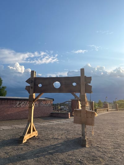 The image depicts an outdoor scene with a wooden stocks structure prominently featured in the foreground. The stocks are constructed from rough-hewn wood, with two circular openings for a person's head and hands, and a pointed arrow shape on one side. A small wooden sign with what appears to be fur trim hangs from a rope attached to the stocks. In the background, a brick wall with cursive lettering in white is visible, along with a metal fence and some streetlights. The ground is covered in gravel. The sky above is a vibrant blue with dramatic, fluffy white clouds and shafts of sunlight. The overall impression is a historical or themed display, possibly part of a park or recreational area in Zhukavichy rural council, Belarus. There are no people visible in the frame.