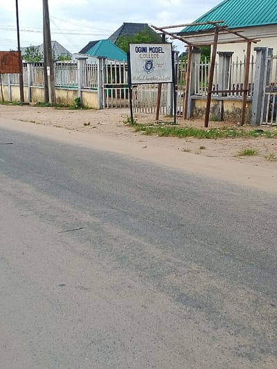 The image shows a signpost for "OGINI MODEL COLLEGE," with a motto: "Motto: Education is Light," in front of a partially built fence along an unpaved road in Ogharefe, Nigeria. The signpost is made of metal and has a white background with blue and black text. There is a small wooden structure with a green roof to the right of the signpost. The building behind the fence is partially visible. The road is dusty, and there are some shrubs and grass in the foreground. There are no people or vehicles in the image. The location is in Ogharefe, Nigeria, at latitude 5.935 and longitude 5.67.