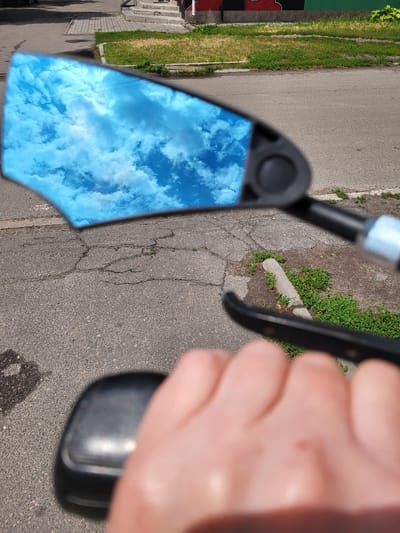 The image depicts a close-up, slightly angled view of a person's hand holding a black object, possibly a camera or a similar device. The object has a reflective surface that captures a bright blue sky filled with fluffy white clouds. The background consists of asphalt pavement with visible cracks and patches of green grass growing through. To the left, a grassy verge with some structures, possibly stairs and a building facade with red and green elements, can be seen. The lighting suggests it is daytime, and the clear sky implies fair weather. The focus is primarily on the reflected sky and the hand holding the object, with the surroundings slightly blurred. There is no visible text in the image. The context of Zolotonosha, Ukraine, places the scene in an urban or suburban outdoor environment.