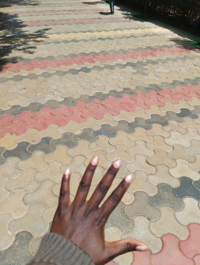 The image depicts an outdoor paved walkway under bright daylight. In the immediate foreground, a dark-skinned left hand is extended, palm facing away, occupying the lower left portion of the frame. The hand features manicured nails with a light, possibly nude or pale pink, polish. The wrist is covered by a ribbed, neutral-toned sleeve.
The walkway, which extends diagonally into the background, is constructed from interlocking concrete pavers arranged in a wavy, tessellated pattern. The pavers are colored in alternating bands of beige, dark grey, and muted reddish-pink. The surface shows minor dirt and wear. Shadows of foliage are distinctly cast onto the left side of the pathway, indicating overhead vegetation.
In the far background, positioned centrally, the lower legs and feet of a person are visible, walking away from the camera. They are clad in dark trousers and shoes. To the right of the paved path, a low, dense green hedge or shrubbery borders the area. The scene is an outdoor environment, described within the context of the city of Hospital ward, Kenya.
