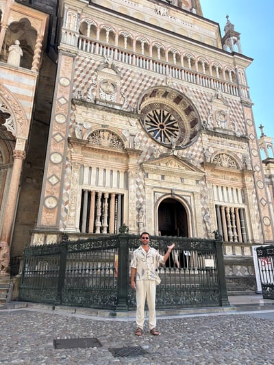A person stands on a cobblestone street in front of a large, ornate building with intricate architectural details. The building, likely a cathedral or similar historical structure, features a rose window, arched doorways, and decorative stonework. The person is wearing a light-colored patterned shirt and pants, sunglasses, and sandals. They are gesturing with their left hand, pointing towards the building. A black wrought-iron gate separates the street from the building's entrance. The sky above is clear and blue, suggesting a bright, sunny day. The time of day appears to be late morning or early afternoon. The location is identified as Mozzo, Italy. No visible text is present in the image.