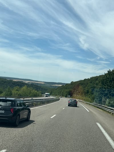 The image depicts a highway scene on a clear day, with a blue sky dotted with wispy clouds. The road curves to the right, bordered by a metal guardrail. Dense green trees line the right side of the road, forming a thick forest that extends up a hillside. On the left side, the terrain slopes downwards, revealing rolling hills covered in a mix of green trees and fields of dry, yellowish vegetation.

Several vehicles are visible on the highway. In the foreground, a black hatchback car is in the left lane, facing away from the camera. Further down the road, a dark-colored car is in the right lane, also moving away. In the distance, a white and green truck is navigating the curve of the highway. The highway itself is paved with asphalt and marked with white lane lines. A road sign is visible on the left side of the road in the distance.

There is no discernible indication in the image that it was taken in Sommedieue, France.