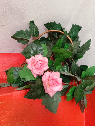 Two artificial pink roses with water droplets on their petals are placed amidst lush green foliage. The flowers and leaves are arranged on a vibrant red surface, which appears to be a ledge or a step. Behind the floral arrangement, a textured off-white wall is visible. The lighting is diffused, casting subtle shadows. The scene is devoid of people or any discernible activity. The location in Tebras, Latvia, is not visually indicated in the image.