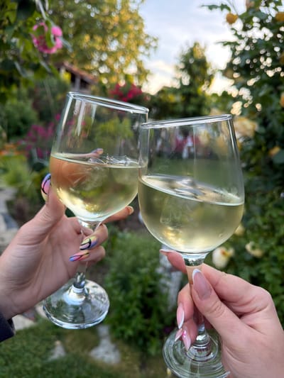 Two wine glasses filled with a pale liquid are being held up in a toast. One hand has a French manicure with a small pink flower on one nail, while the other hand has nails with a colorful, swirled design. The background is a blurred mix of green foliage and flowers, suggesting an outdoor setting. The exact location in Bratia Kunchevi, Bulgaria, is indiscernible from this image.