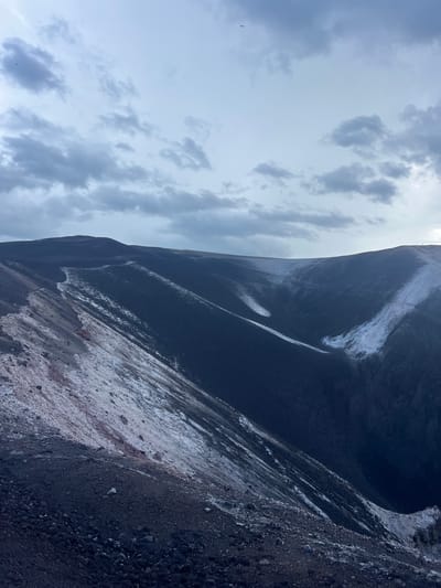 The image shows the rim of a volcanic crater. The ground is covered in dark volcanic rock and ash. Patches of white, possibly snow or mineral deposits, are visible on the slopes. The sky is cloudy and overcast. There are no people or visible man-made structures in the image. The scene is a natural landscape.
