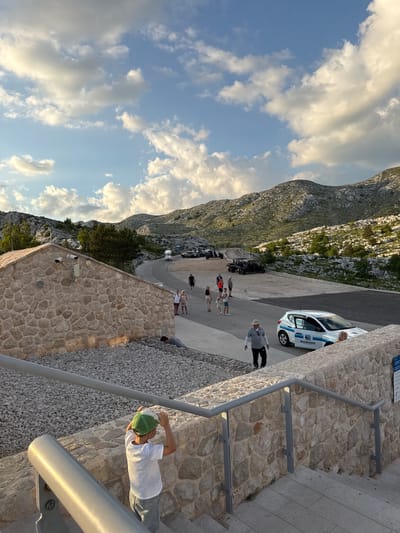 The image depicts a mountainous landscape with a clear sky filled with scattered clouds. In the foreground, a young boy wearing a white t-shirt and grey shorts, with a green cap, stands on stone steps, looking towards the left. A stone building with a tiled roof is visible to the left of the frame, featuring security cameras and vents. Adjacent to the building is an area covered in gravel. Further into the scene, a paved road winds through the terrain, with several off-road vehicles parked and driving. People are seen walking along the road and in the vicinity of the vehicles. A white car with "BIOKOVO" branding is parked on the right side of the road. The background consists of rugged, rocky mountains covered with sparse vegetation. The overall setting suggests an outdoor recreational area or viewpoint in a mountainous region, possibly within Općina Tučepi, Croatia.