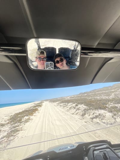 A side-view mirror of a Honda vehicle reflects two people, while the sand dune landscape visible on the side indicates an outdoor location. The blue ocean water is observed through a portion of the image with sky, and the location is likely coastal, which indicates the city of La Capilla, Mexico, may be near a coast.