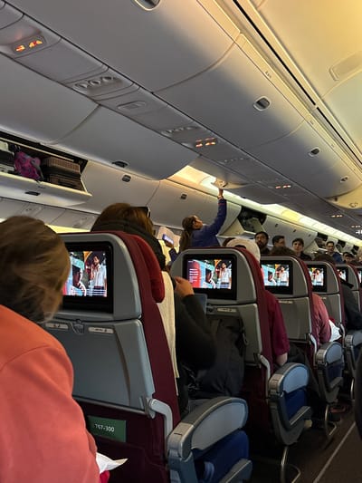 The image captures the interior of an airplane cabin during flight, possibly approaching or departing from Pudahuel, Chile. Rows of burgundy and gray passenger seats are visible, each equipped with personal entertainment screens displaying various media. Occupants of the seats are visible, some watching the screens, others reaching for overhead compartments. The interior lighting is bright. Labels indicate the aircraft type as a Boeing 767-300.