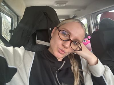 A fair-skinned individual is captured in a headshot selfie from inside a vehicle, located in Menteşe, Turkey. The subject has blonde hair, wears dark brown, oval-framed glasses, and a silver nose ring on their left nostril. They are dressed in a black and off-white/beige zippered jacket with white piping and are looking directly at the camera with puckered lips, their left hand resting casually near their head. The vehicle's interior features a light-colored ceiling and a black garment draped over the headrest directly behind the subject. Through the window to the subject's left, a rocky, possibly snowy, outdoor landscape is visible. To the subject's right, a pink owl-shaped plush toy is positioned on the adjacent seat, with green foliage visible through another window in the background.