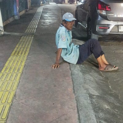A male individual, appearing elderly, is seated on a concrete pedestrian pathway in the Special Capital Region of Jakarta, Indonesia. He wears a light blue collared shirt with a patch on the left sleeve, dark trousers, light brown slide sandals, and a light-colored cap. His hands are positioned on the ground behind him for support, and his knees are bent. A continuous strip of yellow tactile paving is present on the concrete pathway to his left. To his right, the rear section of a grey hatchback vehicle is visible, featuring a license plate beginning with "B," confirming the Jakarta location. The scene is illuminated by artificial light, indicative of nighttime or low-light conditions. A dark building facade is partially visible in the background to the left. The subject's posture suggests a state of rest or waiting.