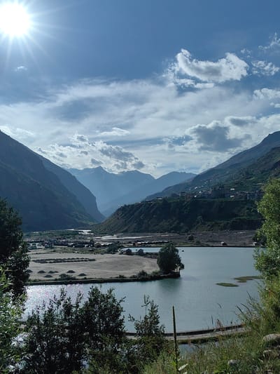 The image captures a serene mountain valley landscape in the vicinity of Yamling, India. The scene is dominated by towering, steep, dark green mountains that flank a wide, sandy riverbed. A calm, bluish-grey body of water, possibly a lake or a wide section of the river, occupies the foreground and mid-ground, reflecting the bright sunlight. The sun itself is a prominent feature in the upper left corner, casting brilliant rays across the sky, which is a clear blue dotted with fluffy white clouds.
On the far side of the water, the landscape transitions to a more settled area with visible signs of human habitation. A small town or village is nestled on the slopes of the mountains, characterized by clusters of buildings with white roofs. The terrain shows evidence of agricultural terracing on the mountainsides. In the mid-ground, along the sandy bank of the river, a collection of small structures, possibly temporary dwellings or storage units, are visible. Several vehicles, including trucks and what appear to be construction equipment, are also present in this area, suggesting some form of activity or development.
The foreground is lush with vegetation, featuring tall grasses and leafy trees, framing the view of the valley and water. The overall impression is one of natural beauty and human presence coexisting within a grand mountainous environment. There are no discernible people in the image, and no specific actions or interactions are captured. The environment is dry and sunlit, with the water providing a contrasting element of calmness.