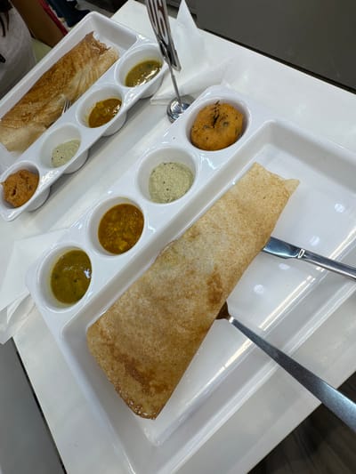A top-down, high-angle shot shows a dosa served on a divided white plastic tray on a white table, likely in a restaurant setting. The dosa, a thin crepe-like pancake, is rolled and positioned lengthwise on the right side of the tray. To its left are four small compartments containing different types of chutney, including a white coconut chutney, a yellow lentil-based chutney, and green chutneys. On a second identical tray towards the bottom of the frame, a smaller dosa is accompanied by three small compartments of similar chutneys. A vada (Indian doughnut) is also visible on the top left tray. Stainless steel utensils, including a fork and a knife, are placed on both trays. Based on the presence of dosas and vadai, it is likely this scene is located in an Indian restaurant. No people are visible, but it appears to be a casual dining setting. While the exact location in the city of Kandy, Sri Lanka, cannot be determined, the presence of South Indian cuisine indicates it might be in an area with a significant Indian population or frequented by tourists.