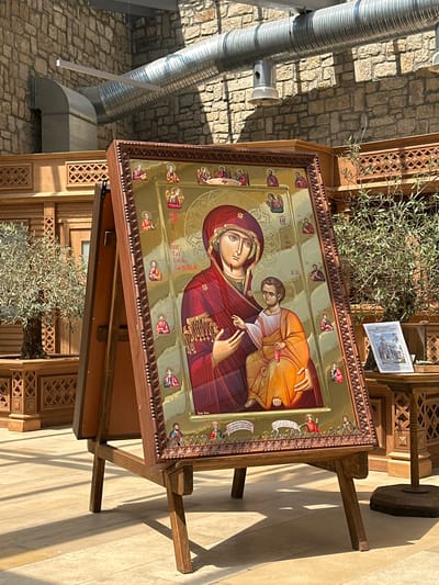 A framed, gilded icon of the Virgin Mary and Child is displayed on a wooden easel in what appears to be an indoor public space with stone walls and wooden architectural elements. The icon is richly decorated with smaller figures and inscriptions. To the right of the icon, a small book or pamphlet with an image of a building is placed on a wooden stand. In the background, there are large metal ventilation ducts running along the stone wall. Green plants in wooden planters are visible on either side of the icon. The floor is tiled, and shadows suggest artificial lighting. This scene is located in the city of Neofit Rilski, Bulgaria.