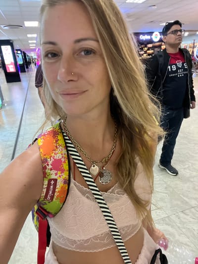 This is a selfie taken in an airport terminal. A woman with blonde hair and a nose ring is in the foreground, wearing a pink lace bralette, a colorful backpack, and several necklaces. Behind her, a man wearing glasses and a black jacket over a red shirt is visible. The background shows an airport environment with shops and lighting. The image appears to have been taken at Bandaranaike International Airport in Katunayake, Sri Lanka, given the presence of a shop labeled "Ceylon Tea."