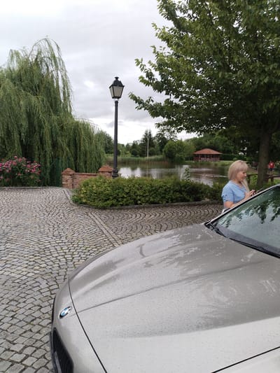 A medium shot captures a silver BMW parked on a cobblestone driveway under an overcast sky, with water droplets visible on the car's hood. A blonde woman in a blue shirt stands near the car, looking at her phone. In the background, a weeping willow tree, a lamppost, and manicured bushes are present. Beyond them, a body of water reflects the sky, and a wooden gazebo with a red roof stands on the opposite shore. The scene is set in Prachovice, Czechia.