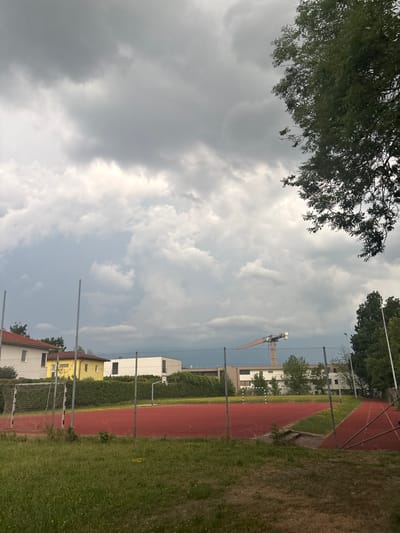 The image depicts an outdoor scene in Gebersdorf, Austria, featuring a red sports court surrounded by a chain-link fence. In the foreground, there is a grassy area with some dry patches and yellow wildflowers. Behind the fence, the sports court is visible, with white lines marking boundaries and a soccer goal on the left side. A running track appears to run along the right side of the sports court.
In the middle ground, there are several buildings. On the left, a house with a red roof is visible. Next to it is a yellow house, and further to the right is a modern white building with dark windows. Behind these structures, a dense green hedge forms a backdrop.
The background is dominated by a cloudy sky, with large, heavy gray clouds suggesting overcast weather. In the distance, a construction crane, painted in yellow and gray, is visible against the sky. Trees, including a large one with dark green foliage on the right edge of the image, frame the scene. The overall impression is that of a sports facility on a cloudy day in a suburban or semi-urban environment.