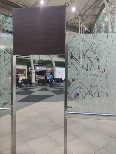 This image captures an indoor public waiting area in Rawa Burung, Indonesia, characterized by modern architecture. In the foreground, a large, blank, dark rectangular display board hangs centrally, partially obstructing the upper view. It is flanked by two frosted glass panels, each adorned with a distinct etched leaf pattern. These panels are supported by polished stainless steel posts and have horizontal handrails at a lower height.

The midground reveals a spacious floor with a tiled pattern, transitioning from light beige squares near the viewer to a checkerboard of dark and light diamond shapes further back. Several rows of black metal bench seating are arranged across this area. On the left, a man in a dark t-shirt and light trousers stands beside a small black suitcase, looking at a mobile phone. To the right, two small commercial stalls are visible: one partially labeled "wehkai" on the left, and a more prominent one on the right named "KOPI HITAM INDONESIA." A person in a purple top is behind the counter of "KOPI HITAM INDONESIA," engaged with items on display. To their right, a cleaning staff member in a blue uniform and gloves holds a cleaning pole or mop, facing away from the center. Reflections of indistinct figures, including one in purple clothing, are visible through the frosted glass panels.

The background features a high, vaulted ceiling structure with exposed white metal trusses and large white fabric or panel sections. Bright, square-shaped lights are embedded in the ceiling. Additional glass panels in the upper structure suggest natural light entry. Greenery, possibly hanging plants, is visible along a ledge above the commercial stalls. A decorative wall panel with a colorful geometric pattern is present on the far right.