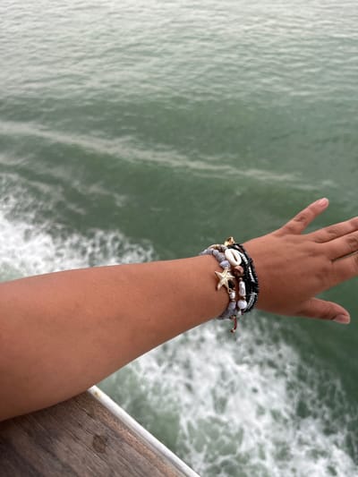 A dark-skinned arm and hand with multiple bracelets is positioned in the upper right quadrant of the image against a background of rippled green water. The arm, extending from the bottom of the frame to above the wrist, is adorned with multiple beaded bracelets, one with a small gold starfish charm and another with a seashell. The fingers of the hand are extended. The water in the background appears turbulent with foamy areas, suggesting movement. A corner of a pier is visible in the bottom right. There is no information to support it being taken in Loon-Plage, France.